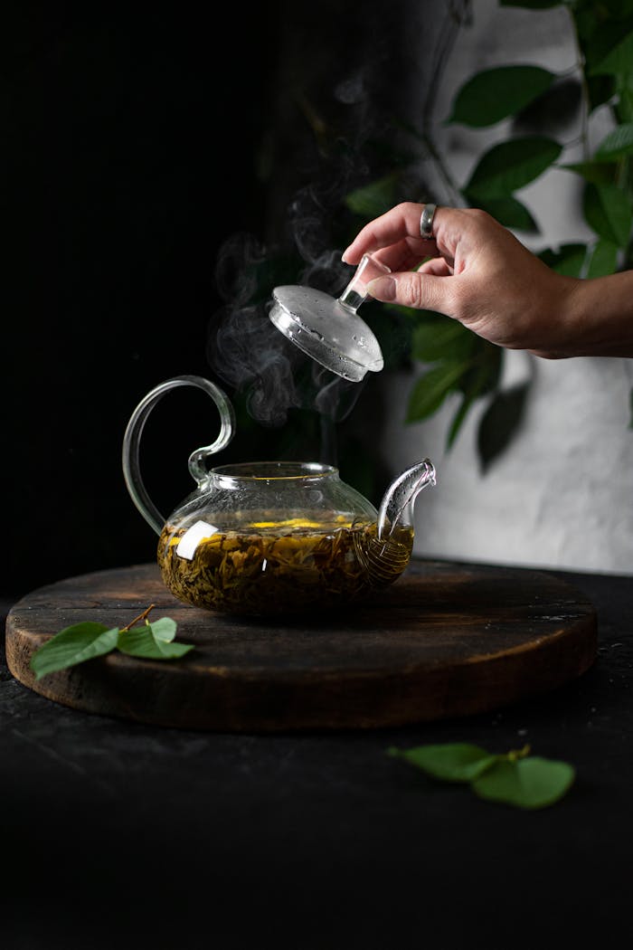 Glass teapot with aromatic green tea, steam rising, hand lifting lid, natural setting.